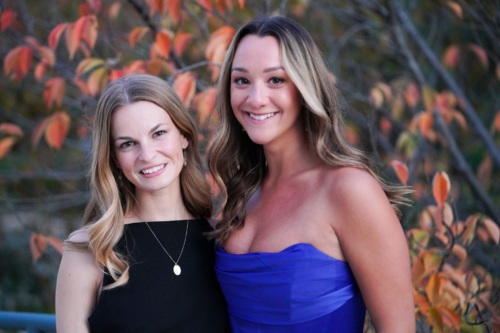 Two women smile in front of fall leaves at an annual gala