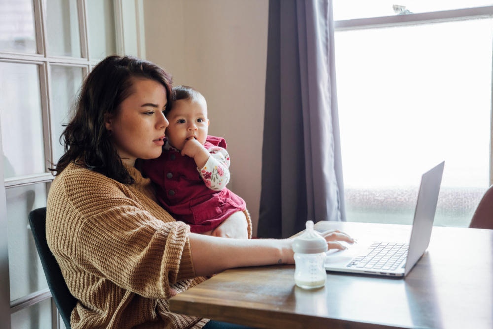 Mom holding baby and working from home on laptop at dining room table