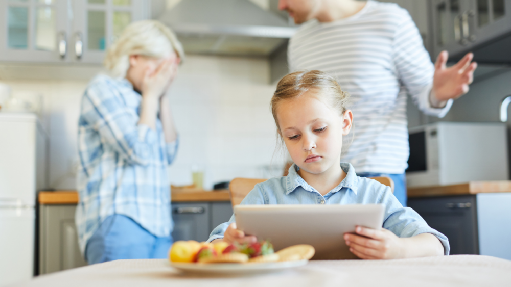 A young girl looks at a tablet while her parents are visibly frustrated in the background