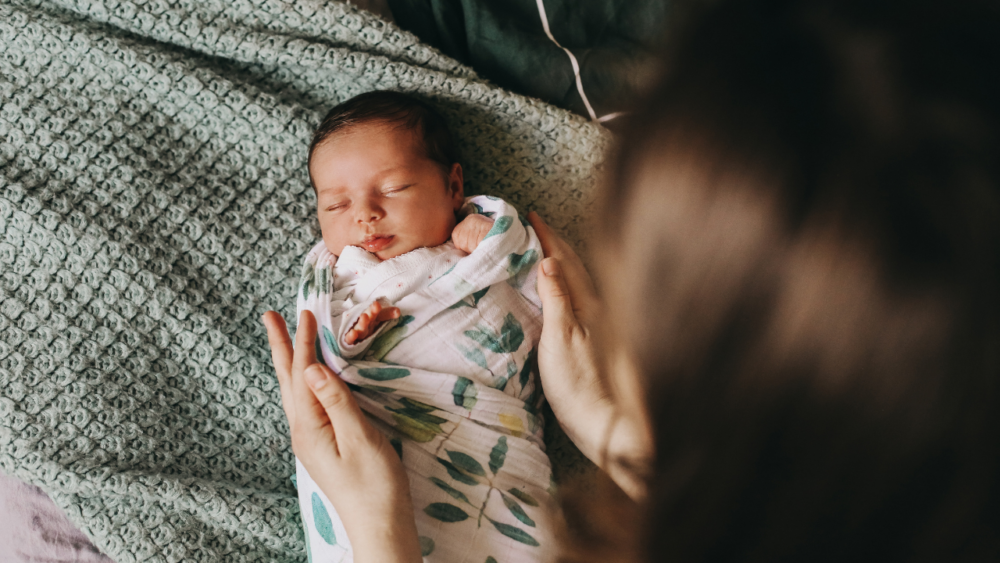 A woman looks on as a young baby sleeps in a swaddle