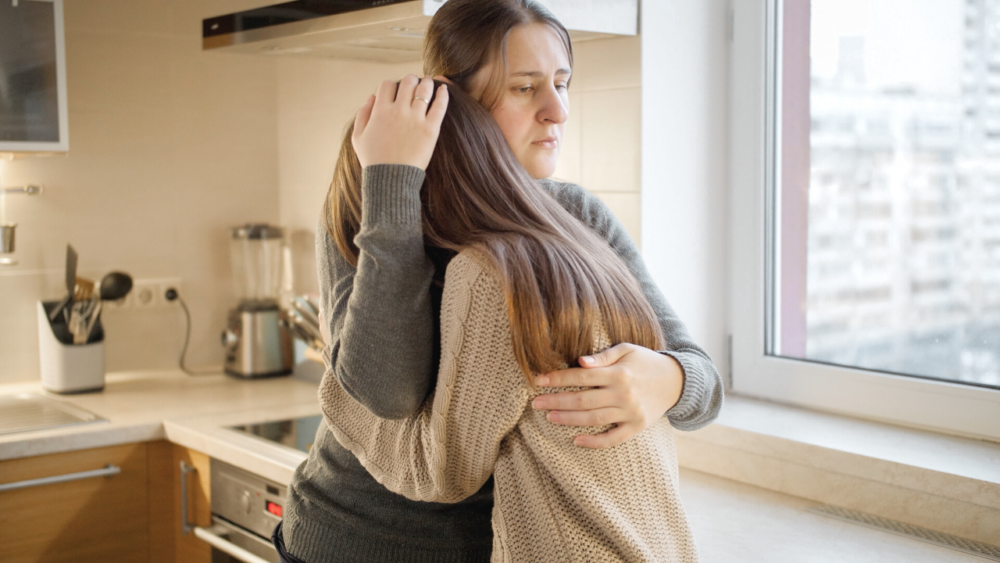 A woman hugs her daughter with a forlorn look on her face