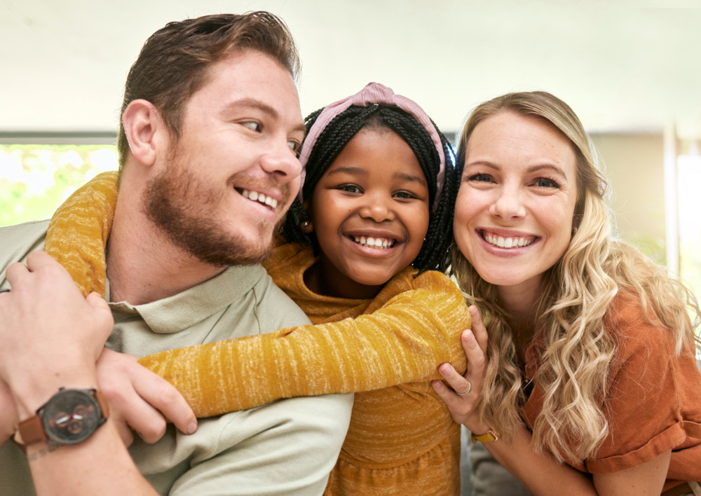 An interracial family with three members - a mother, father, and daughter smile and hug while looking at the camera. Photo for national adoption awareness month training!